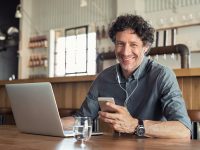Portrait of businessman using smartphone with earphones and laptop for a business conference. Cheerful smiling business man sitting in restaurant listening to music. Happy man enjoying free wireless internet connection at coffee shop.